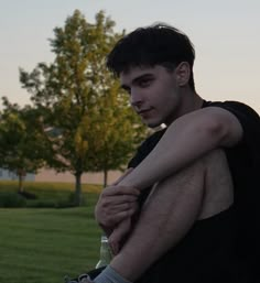 a young man sitting on the ground with his arm around someone's leg, in front of some trees