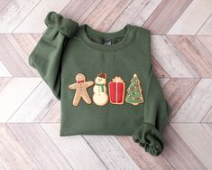 a green sweater with ginger, snowman, and christmas cookies on the front is laying on a wood floor