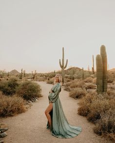 a woman in a long dress standing on a dirt road next to cacti