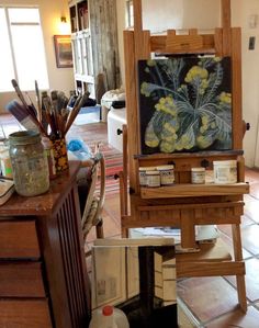 an easel sitting on top of a wooden floor next to a table with paint and brushes