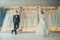 a man and woman standing in front of wedding gowns hanging on the wall behind them