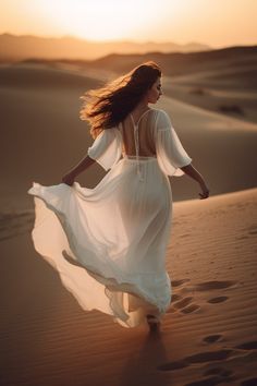 a woman in a white dress is walking through the sand dunes at sunset with her hair blowing in the wind