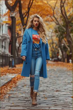 long blonde beachy waves, flamboyant natural kibbe body type, bright blue wool coat, graphic t-shirt with bold red and yellow print, high-waisted distressed skinny jeans, tan ankle boots, large statement earrings, casual yet stylish autumn outfit with a mix of vibrant colors and modern streetwear elements. Bright Outfits Casual, Bright Blue Jacket Outfit, Light Blue Jeans Outfit Fall, Blue Jeans Outfit Fall, Natural Kibbe Body Type, Flamboyant Natural Kibbe, Streetwear Layering, Light Blue Jeans Outfit, Natural Kibbe