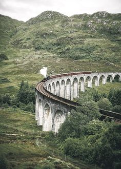 a train traveling over a bridge on top of a lush green hillside with mountains in the background