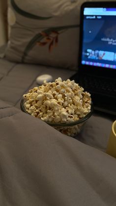 a glass bowl filled with popcorn sitting next to a laptop computer on top of a bed