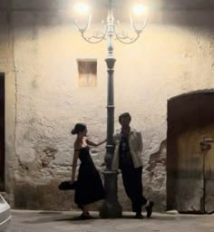 a man and woman standing next to a lamp post in an alleyway at night