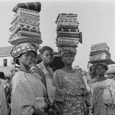 black and white photograph of three women carrying stacks of bags on their heads, with other people standing around