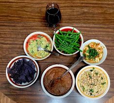 a wooden table topped with white bowls filled with different types of food next to a glass of wine