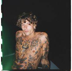 a man with tattoos on his body sitting in front of a green light and looking at the camera