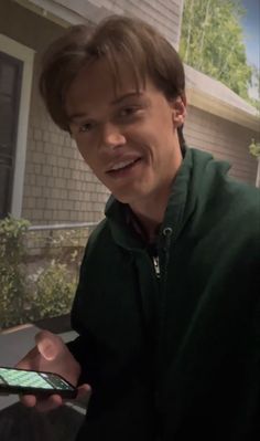 a young man holding a smart phone in his hand and smiling at the camera while standing outside