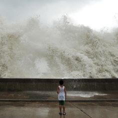 a person standing in front of a large wave crashing over the sea wall on a cloudy day