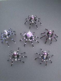 six spider brooches are shown on a gray surface, each with purple and white beads