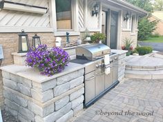 an outdoor grill with purple flowers on the counter next to a brick patio and house