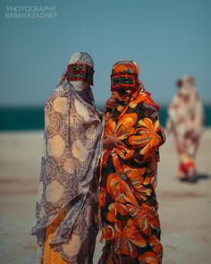 two women in colorful clothing standing next to each other on the beach with people walking behind them