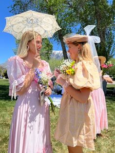 two women standing under an umbrella talking to each other on the grass in front of some trees