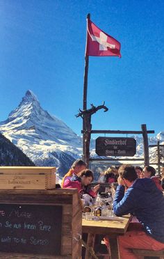 some people are sitting at a table in front of a mountain with a swiss flag