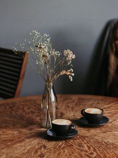 two cups and saucers sitting on a table with flowers in a vase next to them