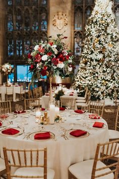 a table set up with white linens, red napkins and gold chairs in front of a christmas tree