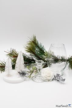 white flowers and pine branches are sitting in front of wine glasses on a white background