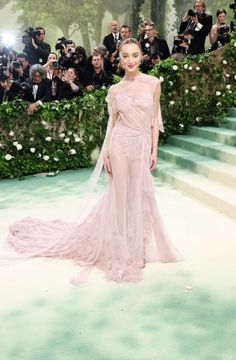 a woman in a pink gown stands on the green carpet with photographers taking pictures behind her