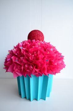 a blue cupcake with pink flowers in it on a white tablecloth and a red ornament hanging from the top