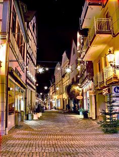 an empty cobblestone street at night with lights strung from the buildings on both sides