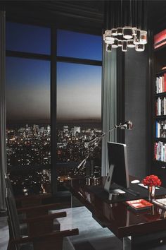 a room with a desk, bookshelf and large window overlooking the city at night