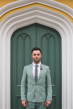 a man standing in front of a green door wearing a suit and tie with his hands in his pockets