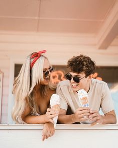 two people sitting on a ledge with ice cream cones in their hands and looking at each other