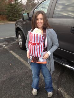 a woman holding a bag full of popcorn standing in front of a parked car and smiling at the camera