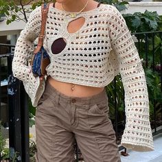 a woman standing in front of a fence wearing brown pants and a white crochet top