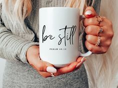 a woman holding a coffee mug with the word be still on it in her hands