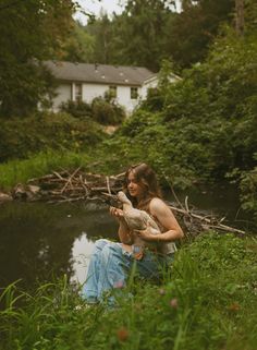 a woman sitting in the grass holding a bird next to a body of water with a house in the background