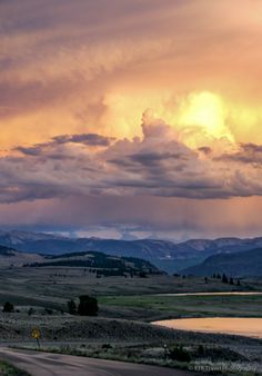 the sun is setting over an open field with mountains in the distance and clouds above