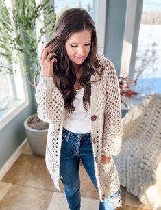 a woman standing in front of a window wearing ripped jeans and a white top with an open knit cardigan