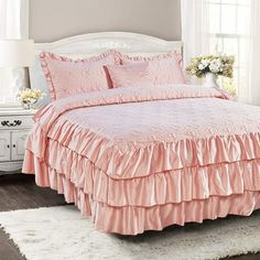 a pink bed with ruffled bedspread and pillows in a white room next to a window