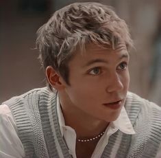 a young man with short hair wearing a sweater and pearl necklace looking at the camera