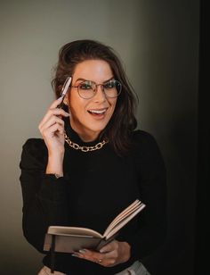 a woman wearing glasses holding a book and talking on a cell phone while smiling at the camera