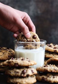 a person picking up a cookie from a glass of milk with chocolate chip cookies in the background