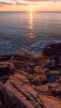 the sun is setting over the ocean with rocks in front of it and water below