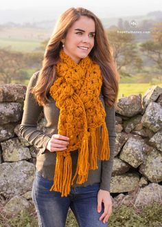 a woman standing in front of a stone wall wearing a mustard colored crochet scarf