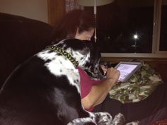 a woman sitting on a couch with her dog and looking at an electronic device in her lap