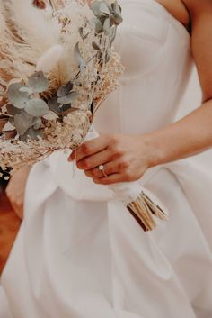 a woman in a white wedding dress holding a bridal bouquet with greenery on it