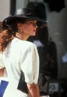 a woman in a white dress and black hat walking down the street with her hand on her hip