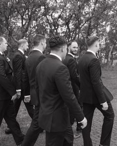 a group of men in suits walking across a grass covered field with trees behind them