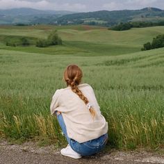 a woman sitting on the side of a road next to a lush green field with hills in the background