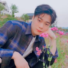 a young man sitting in the grass next to pink flowers and holding his hand on his chin