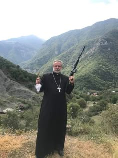 a priest holding two large metal objects in front of mountains and valleys with hills behind him