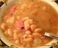 a white bowl filled with beans and ham on top of a floral table cloth next to a spoon