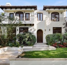 a large white house with lots of windows and plants in the front yard, on a sunny day